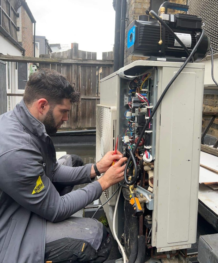 A maintenance technician kneeling beside an open HVAC or air conditioning unit, inspecting and adjusting wiring and components with a screwdriver.