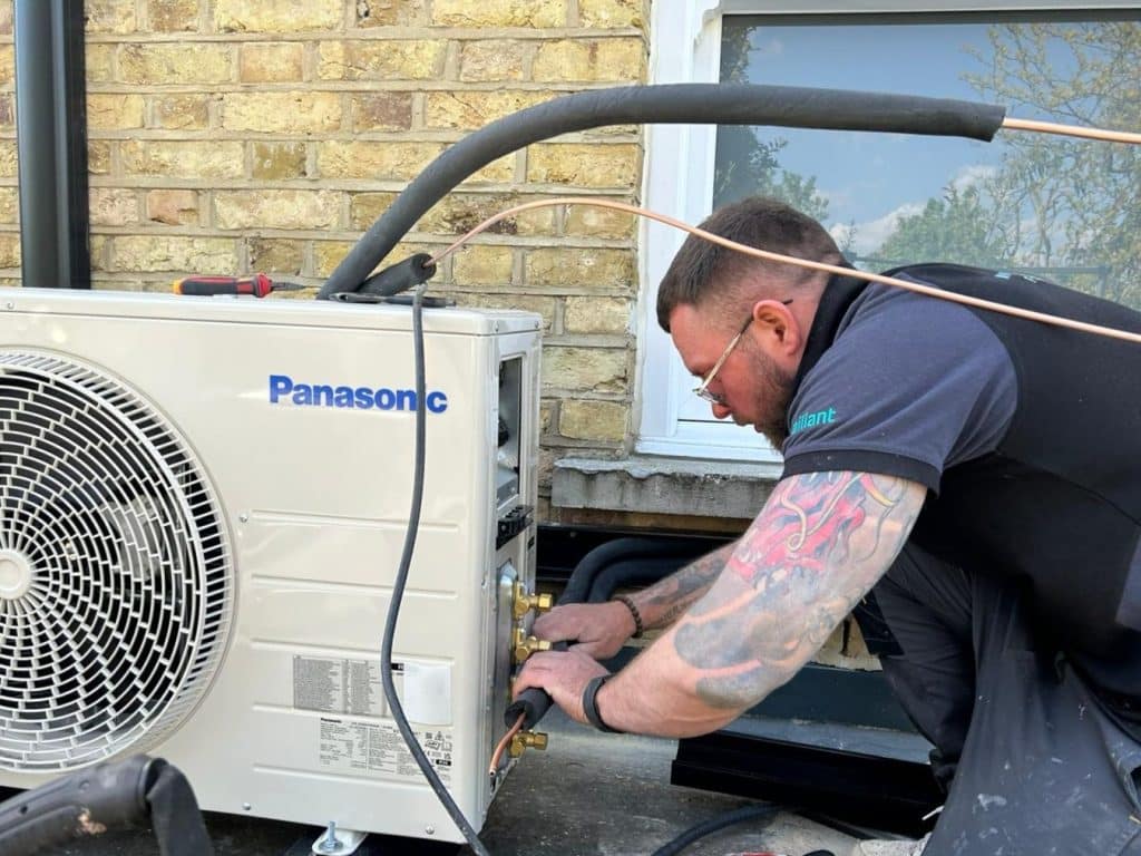 Engineer servicing an outdoor Panasonic air conditioning unit beside a brick wall